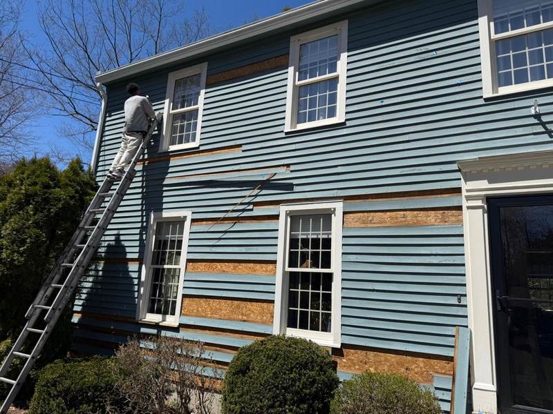 A painter on a ladder at a blue house where sections of siding are being replaced.