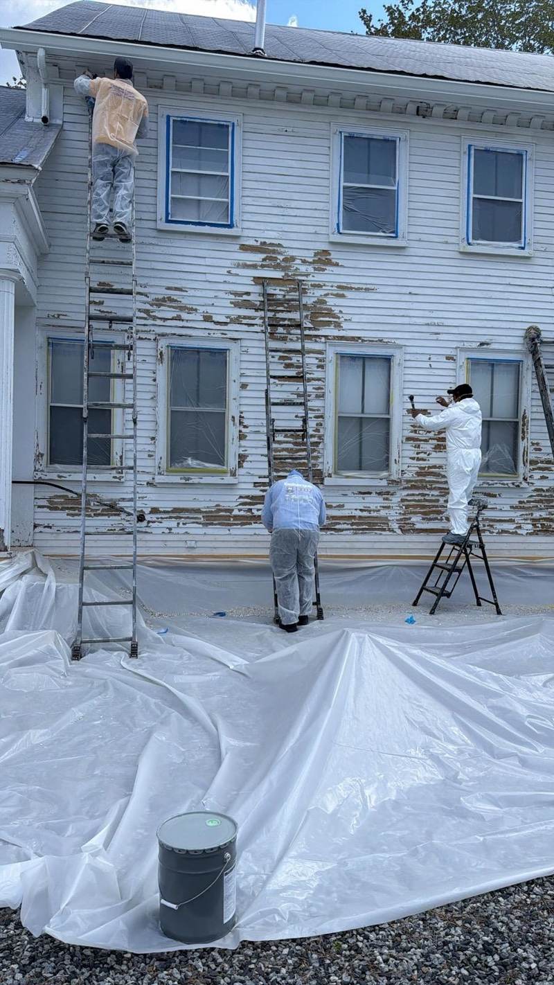 Two painters working on a white house with drop cloths spread across the ground.