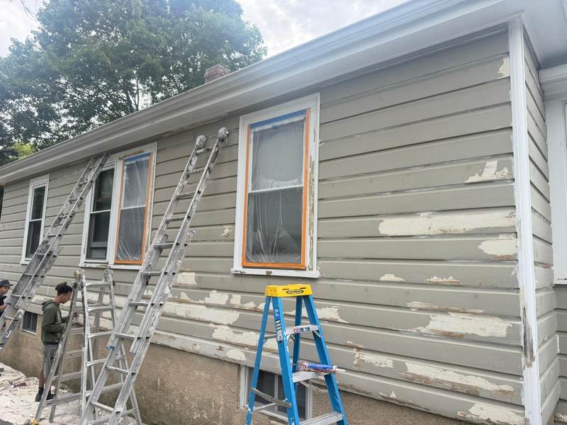Ladders set against a gray-green wall being scraped, with the windows masked for prep.