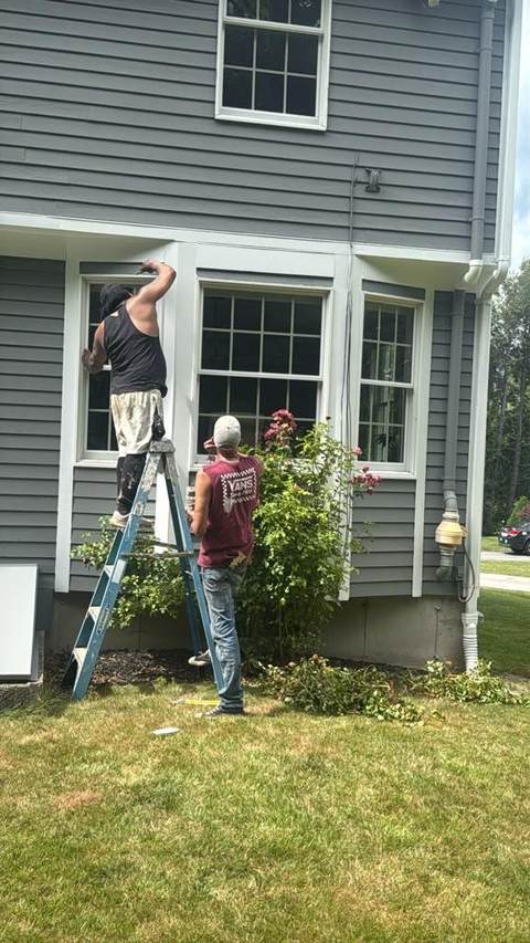 Two painters at a ladder working on the window trim of a gray house.