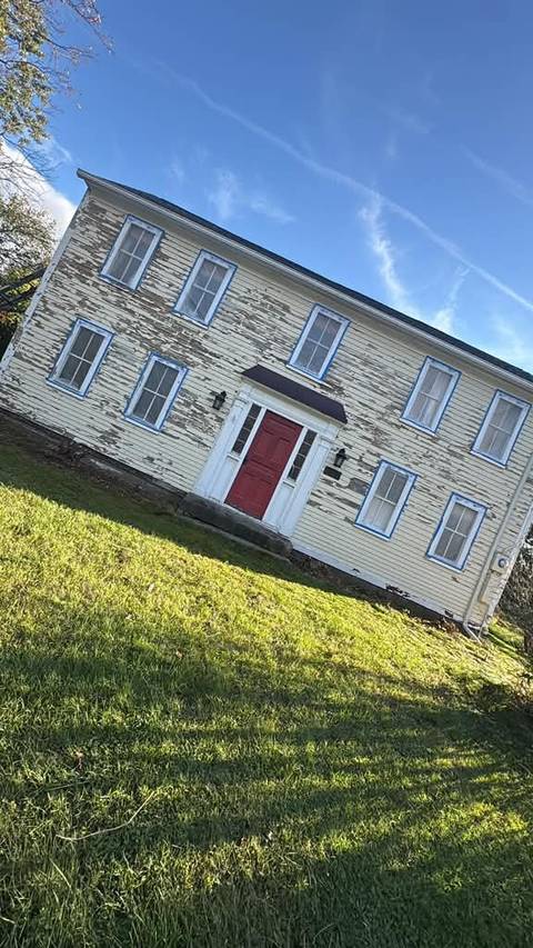 The pale yellow colonial with peeling paint and a red door, seen from the lawn.