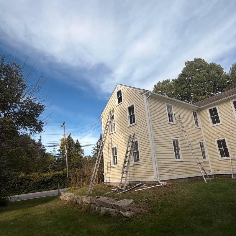 Ladders set against a pale yellow colonial during exterior preparation.