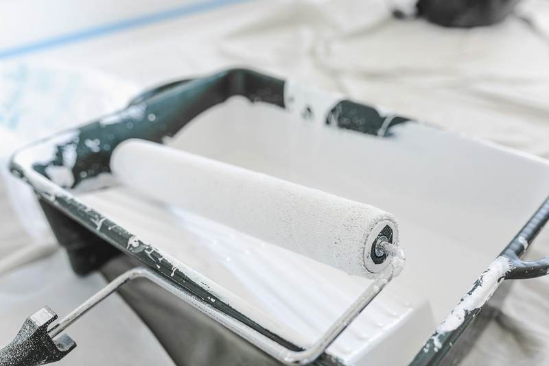 A roller resting in a tray of white paint on a covered floor. Reference photograph.
